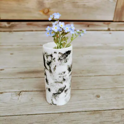 The Rustic Elegance Porcelain Mini Vase, designed in white with black patterns, holds a bunch of tiny blue flowers on a wooden surface.
