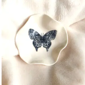 A white ceramic Butterfly Ring Holder (7cm x 7cm) with irregular edges and a black butterfly illustration in the center is placed on a soft white fabric.