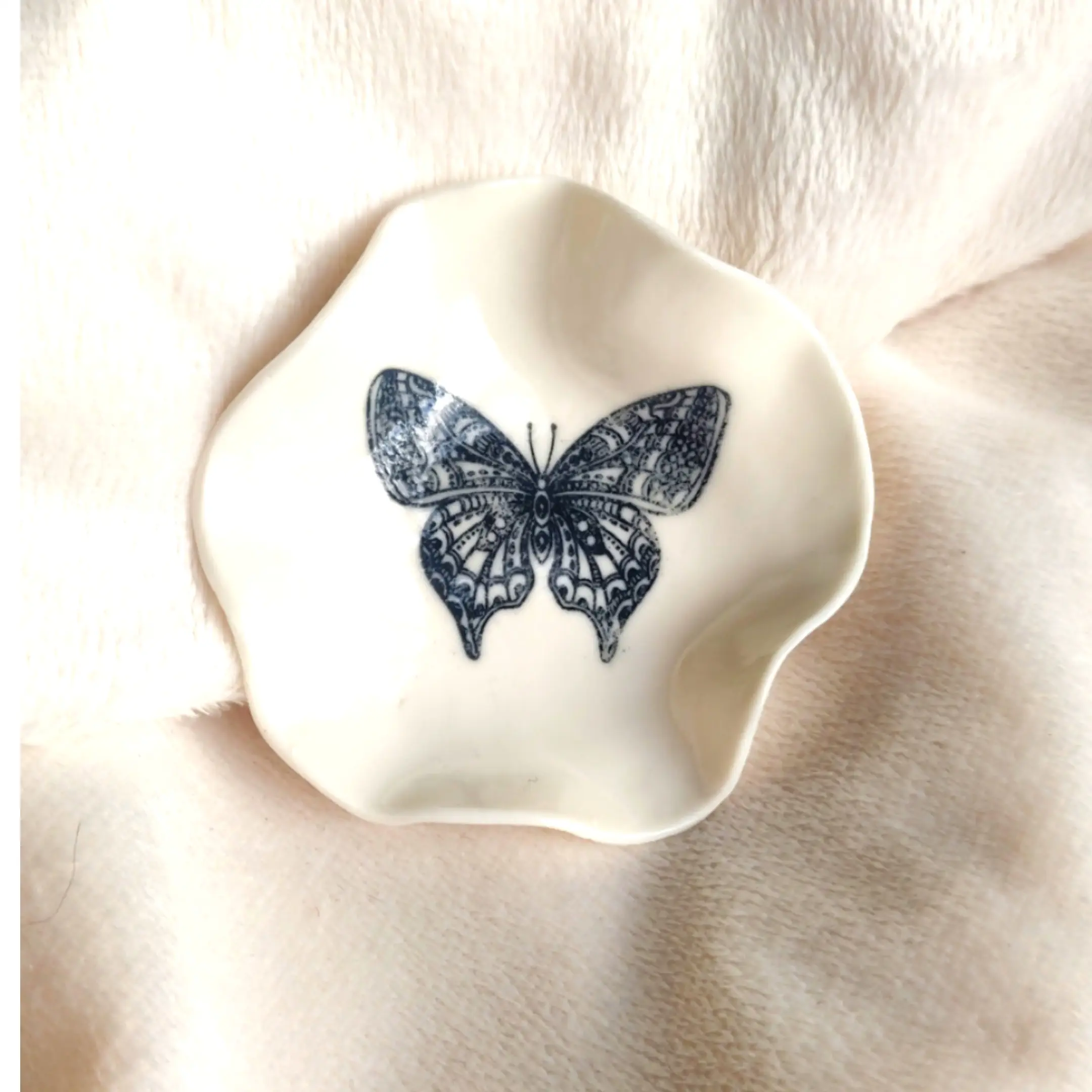 A white ceramic Butterfly Ring Holder (7cm x 7cm) with irregular edges and a black butterfly illustration in the center is placed on a soft white fabric.