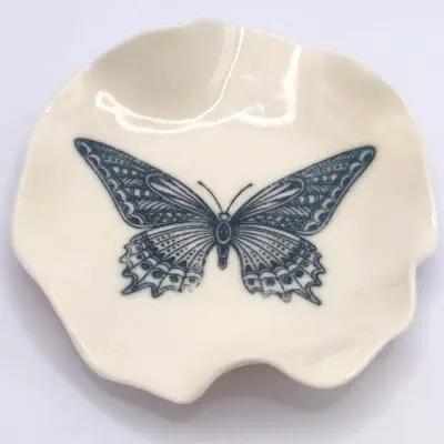 A small, cream-colored ceramic dish with a detailed black butterfly illustration at the center.
