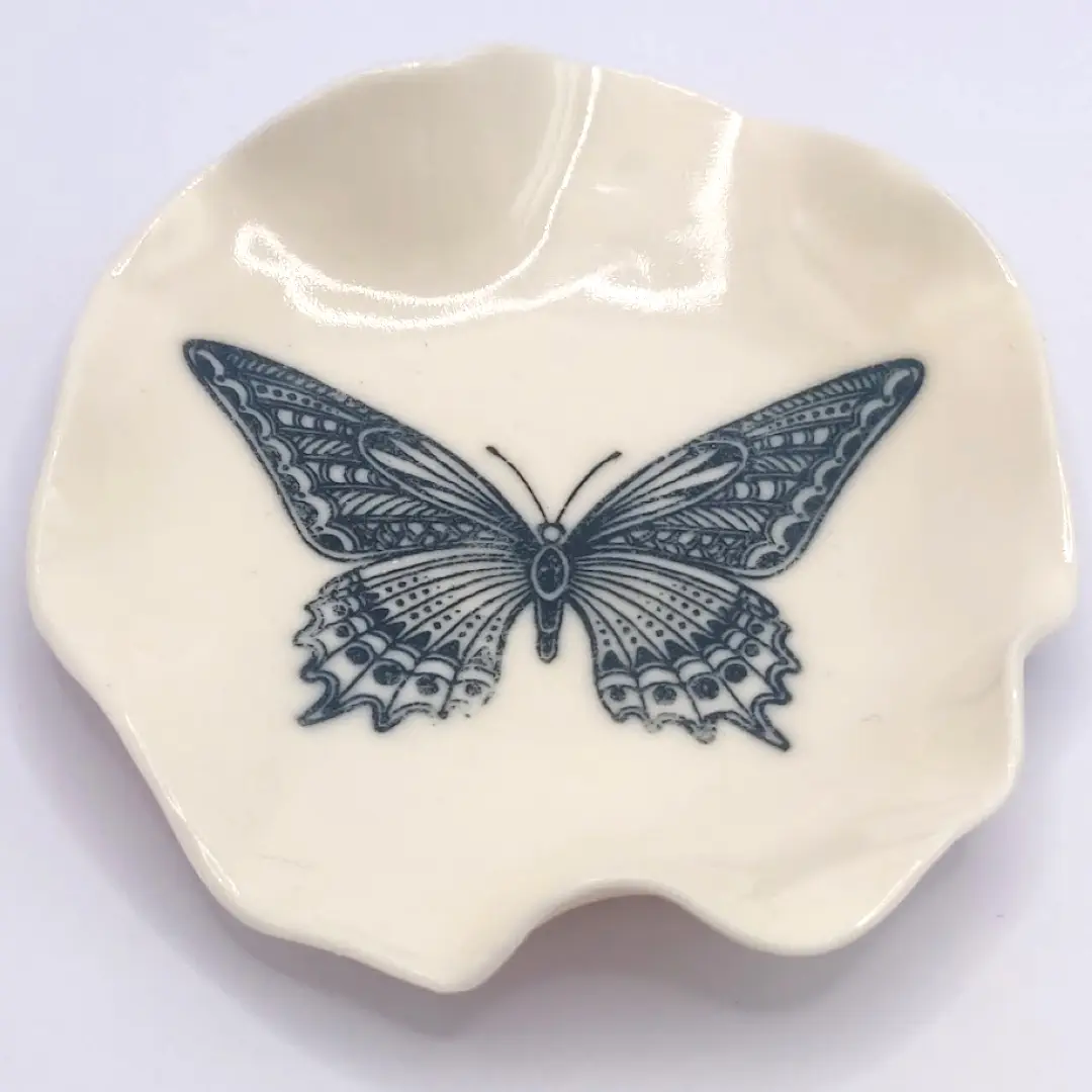 A small, cream-colored ceramic dish with a detailed black butterfly illustration at the center.
