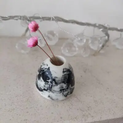 Ethereal Swirl Miniature Vase in black and white marble design with two pink dried flowers, set on a light surface adorned with clear string lights in the background.