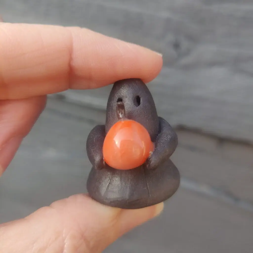 A hand holds a small, dark figurine known as the Midnight Mini Ghost Figurine 🎃, shaped like a bird with an orange sphere in its grasp.