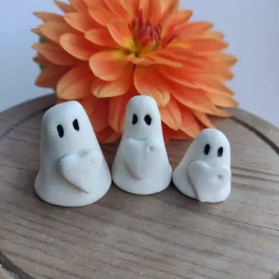 The Love Ghost Trio, featuring three small white clay ghost figures with black eyes and heart details, is showcased in front of an orange flower on a wooden surface.