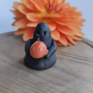 The Midnight Mini Ghost Figurine 🎃, a small black clay figure holding an orange pumpkin, is placed on a wooden surface with a large orange flower in the background.