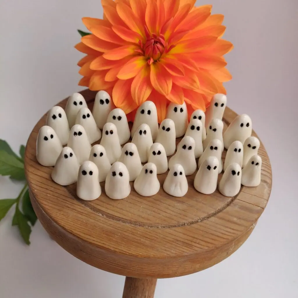 The Mini Ghostly Gathering Porcelain Collection, featuring small white ghosts with black eyes, is arranged on a wooden stool with an orange dahlia flower in the background.
