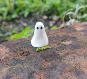 A piece from the Mini Ghostly Gathering Porcelain Collection, featuring a small, smooth white figurine with two black dots for eyes, is displayed on a moss-covered brick outdoors.