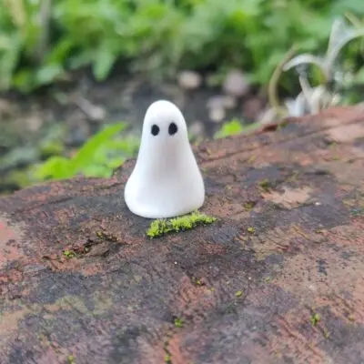 A piece from the Mini Ghostly Gathering Porcelain Collection, featuring a small, smooth white figurine with two black dots for eyes, is displayed on a moss-covered brick outdoors.
