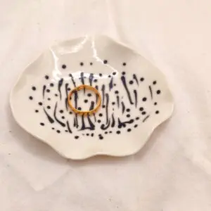 A gold ring rests on the Squiggles and Dots porcelain ring holder, featuring black dotted and striped patterns, placed on a white fabric background.