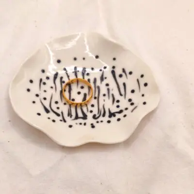 A gold ring rests on the Squiggles and Dots porcelain ring holder, featuring black dotted and striped patterns, placed on a white fabric background.