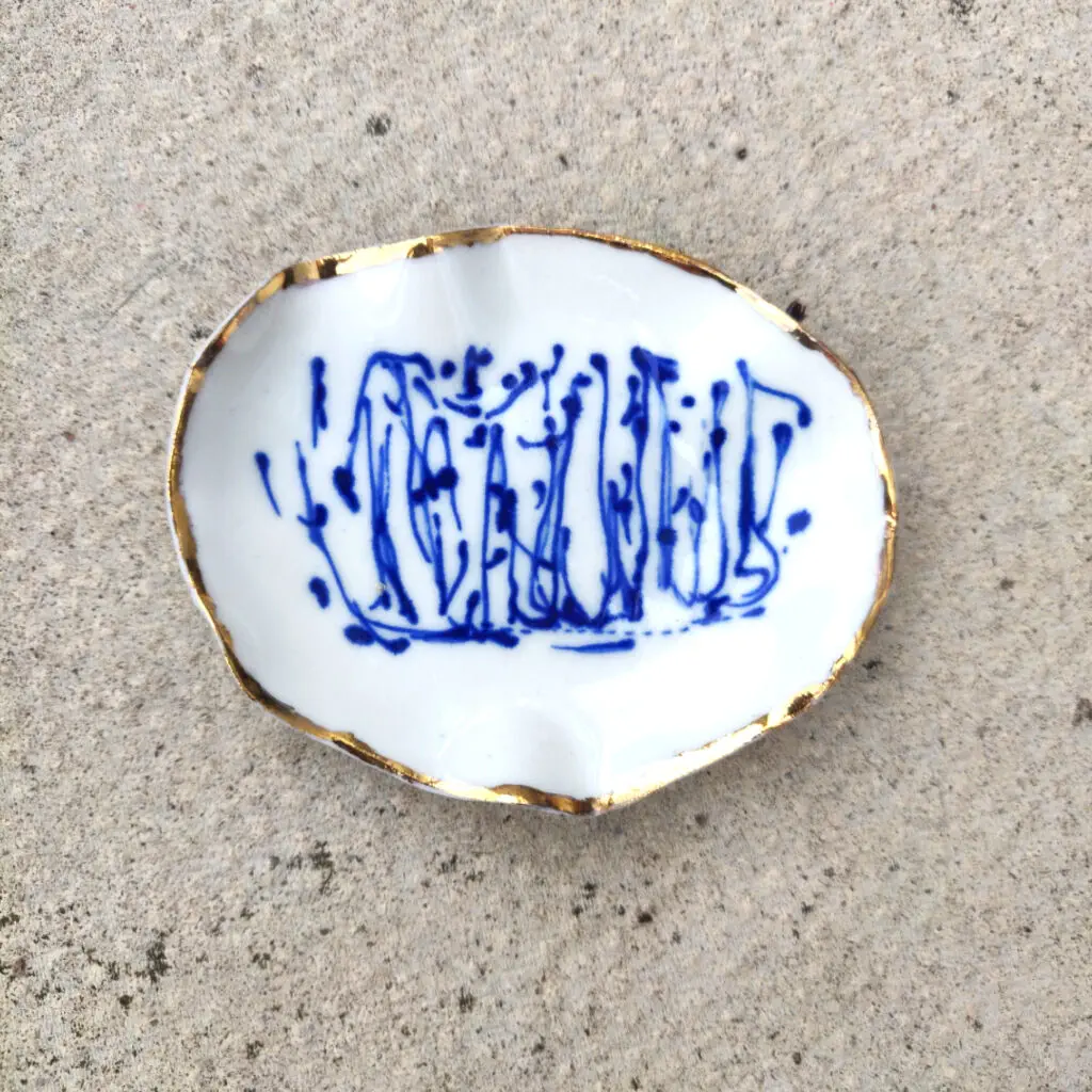 The Blue and White Porcelain Ring Holder with gold lustre features a white base adorned with abstract blue patterns and is elegantly finished with gold-trimmed edges, all set against a textured gray surface.