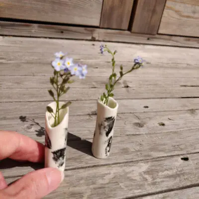 Two Ink-Stroke Miniature Porcelain Vases, featuring black markings on a white cylindrical surface and each containing delicate purple flowers, rest on a wooden surface as a hand carefully adjusts one of them.