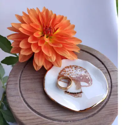 An orange flower is positioned beside the Enchanted Forest Porcelain Mushroom Ring Holder/Trinket with Gold Lustre, which holds two gold rings and rests on a round wooden surface.