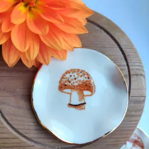 An orange flower is set beside the Enchanted Forest Porcelain Mushroom Ring Holder/Trinket with Gold Lustre, elegantly placed on a wooden surface.