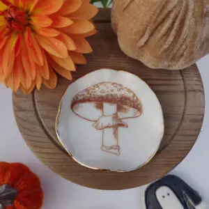 Ceramic dish with a mushroom illustration, surrounded by a dahlia flower, fabric pumpkin, and wooden circular board.