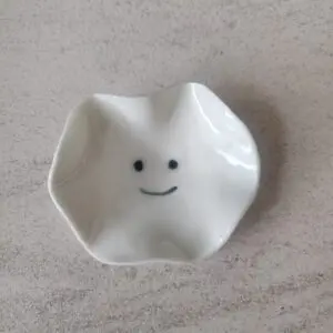 A compact, white ceramic dish featuring a wavy edge and adorned with a minimalist smiley face design at its base, known as the Mini Smiley Face.