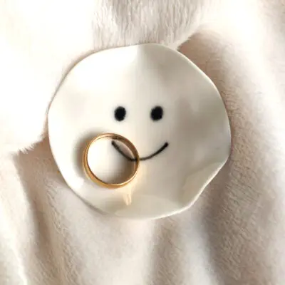 The Smiling Face Ring Holder, a small ceramic dish with a drawn smiley face, holds a gold ring and is placed on a soft, light-colored fabric.