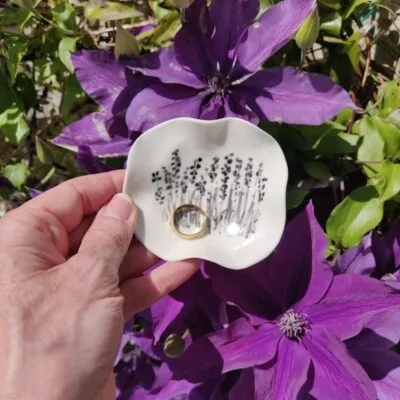 A hand holding the Enchanted Meadow Porcelain Ring Holder, adorned with a black floral pattern and a gold ring, set against large purple flowers in the background.
